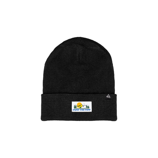 A black knit beanie with a folded cuff featuring a patch with the phrase "ENJOY THE NOW" in bold lettering, accompanied by a graphical element of a mountain and sun.