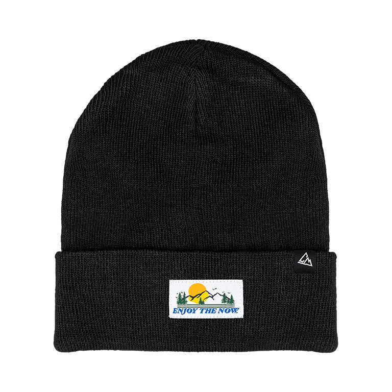 A black knit beanie with a folded cuff featuring a patch with the phrase "ENJOY THE NOW" in bold lettering, accompanied by a graphical element of a mountain and sun.