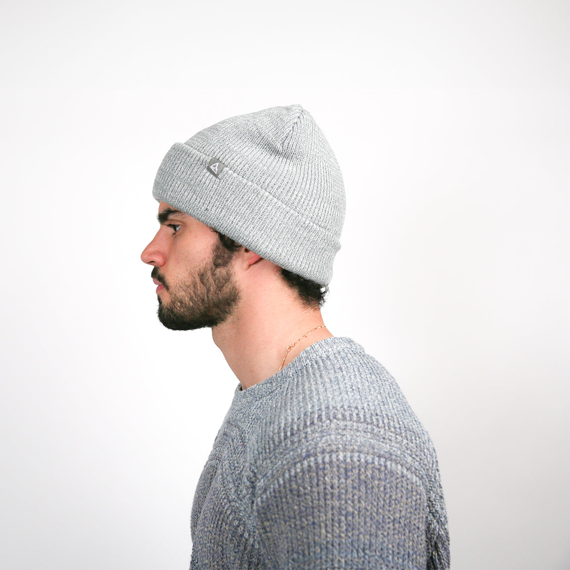 Viewed from the side, the light grey beanie reveals a fine ribbed pattern, along with a small, dark logo patch, contributing to the hat’s understated style.