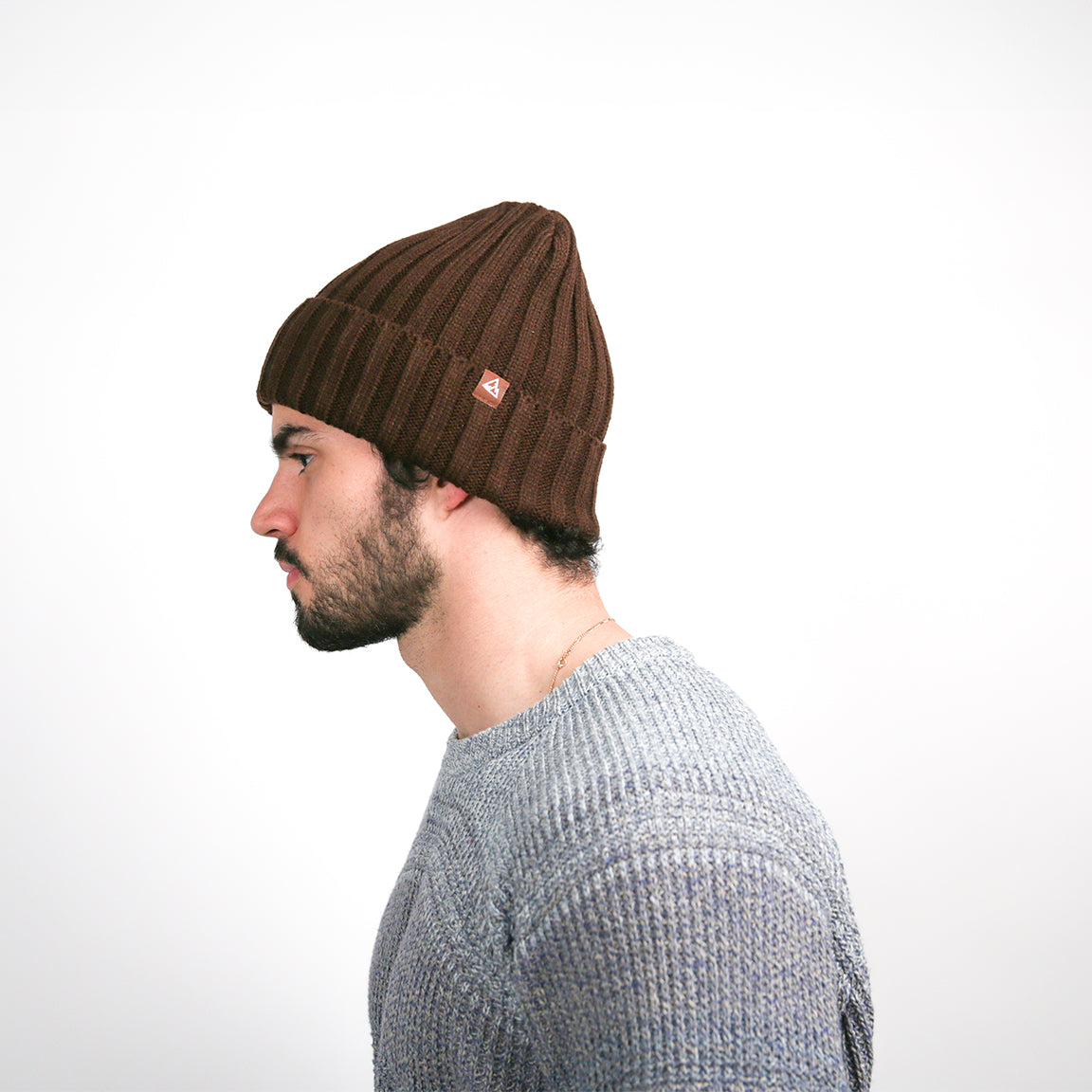 A side profile view, of the dark brown beanie is shown. The ribbed texture and the tan logo are now visible from the side. The beanie covers the ears and has a relaxed fit, with the material gathering slightly at the top for a casual look. The texture of the grey knit sweater is also apparent from this angle.
