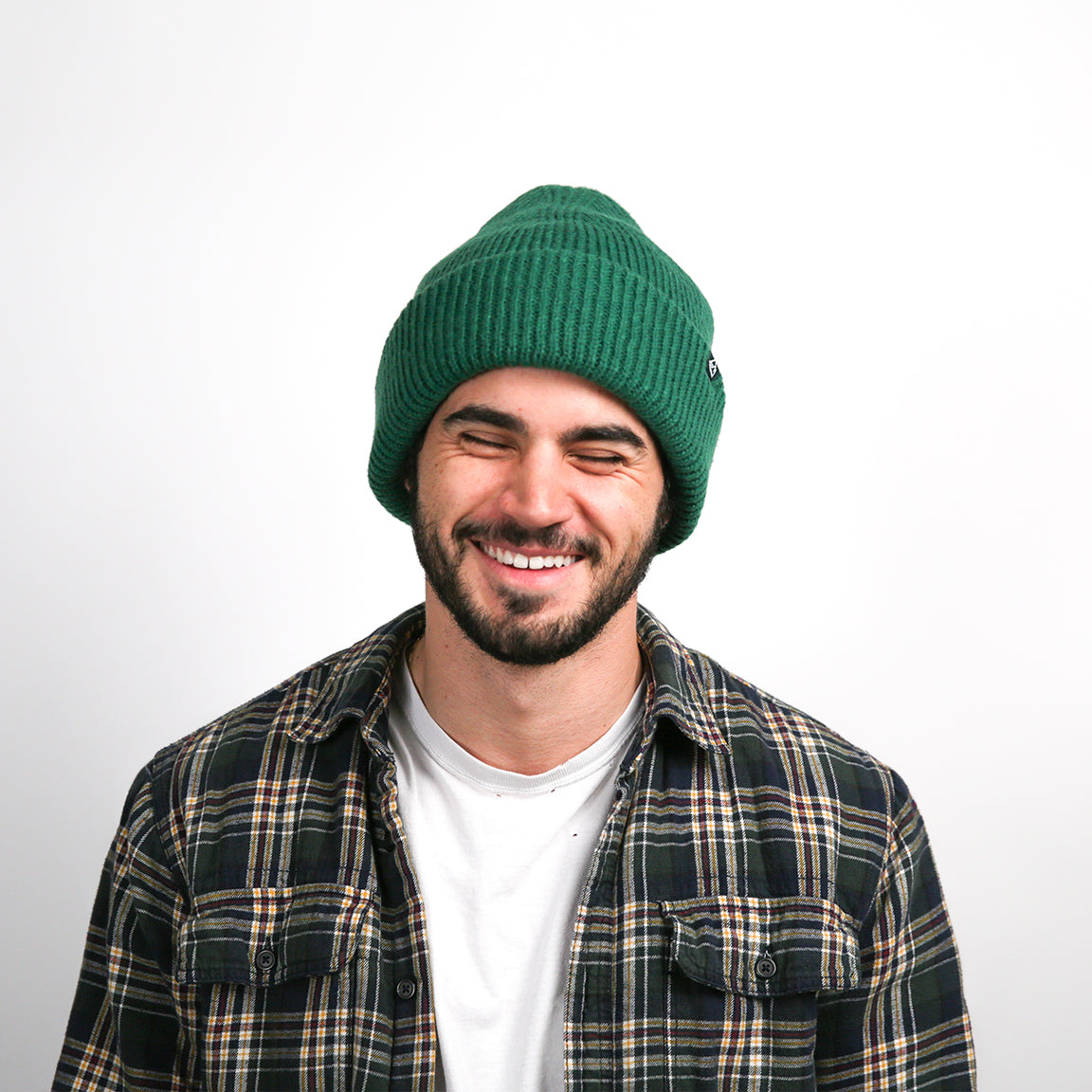 A person is smiling while wearing a ribbed knit beanie in a deep forest green shade. The beanie has a relaxed fit with a rolled-up brim, providing a casual and cozy style.