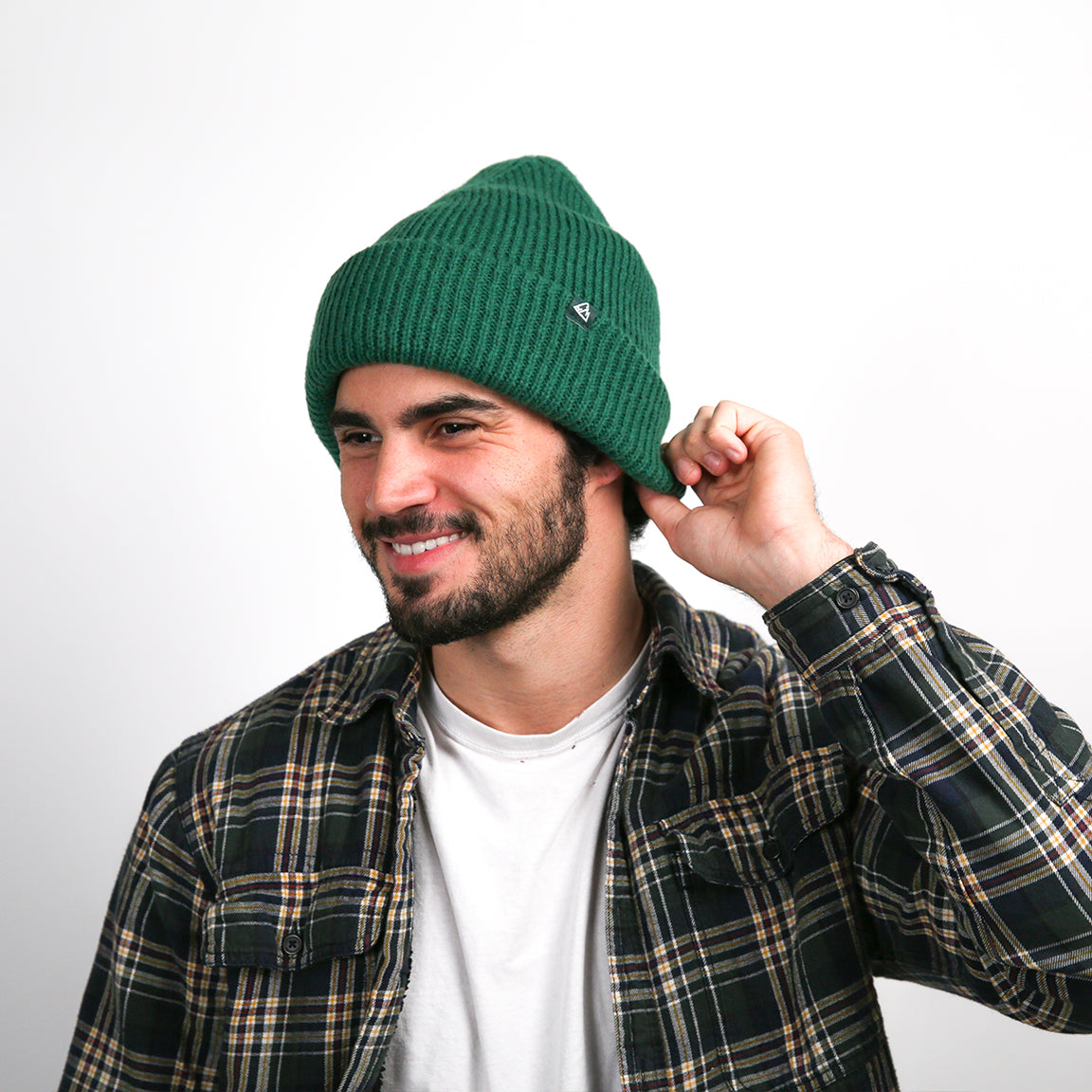 A person is smiling while wearing a ribbed knit beanie in a deep forest green shade. The beanie has a relaxed fit with a rolled-up brim, providing a casual and cozy style.