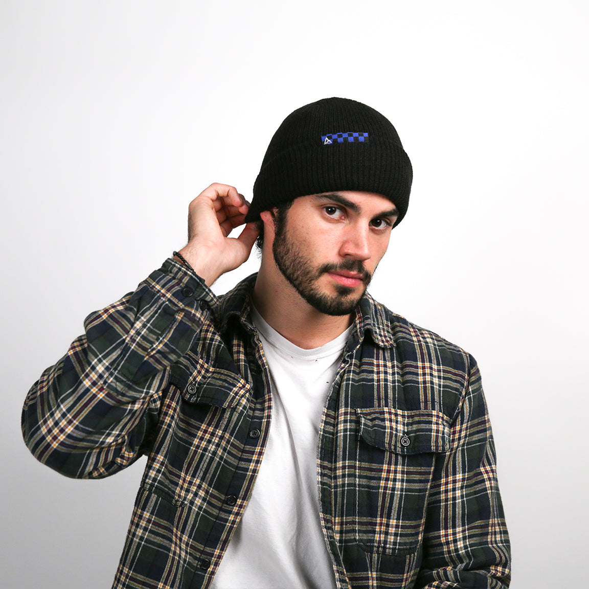A person is adjusting a black ribbed knit beanie that has a rolled-up brim and features a small blue checkered logo patch on the side, accentuating the hat's snug fit and classic style.