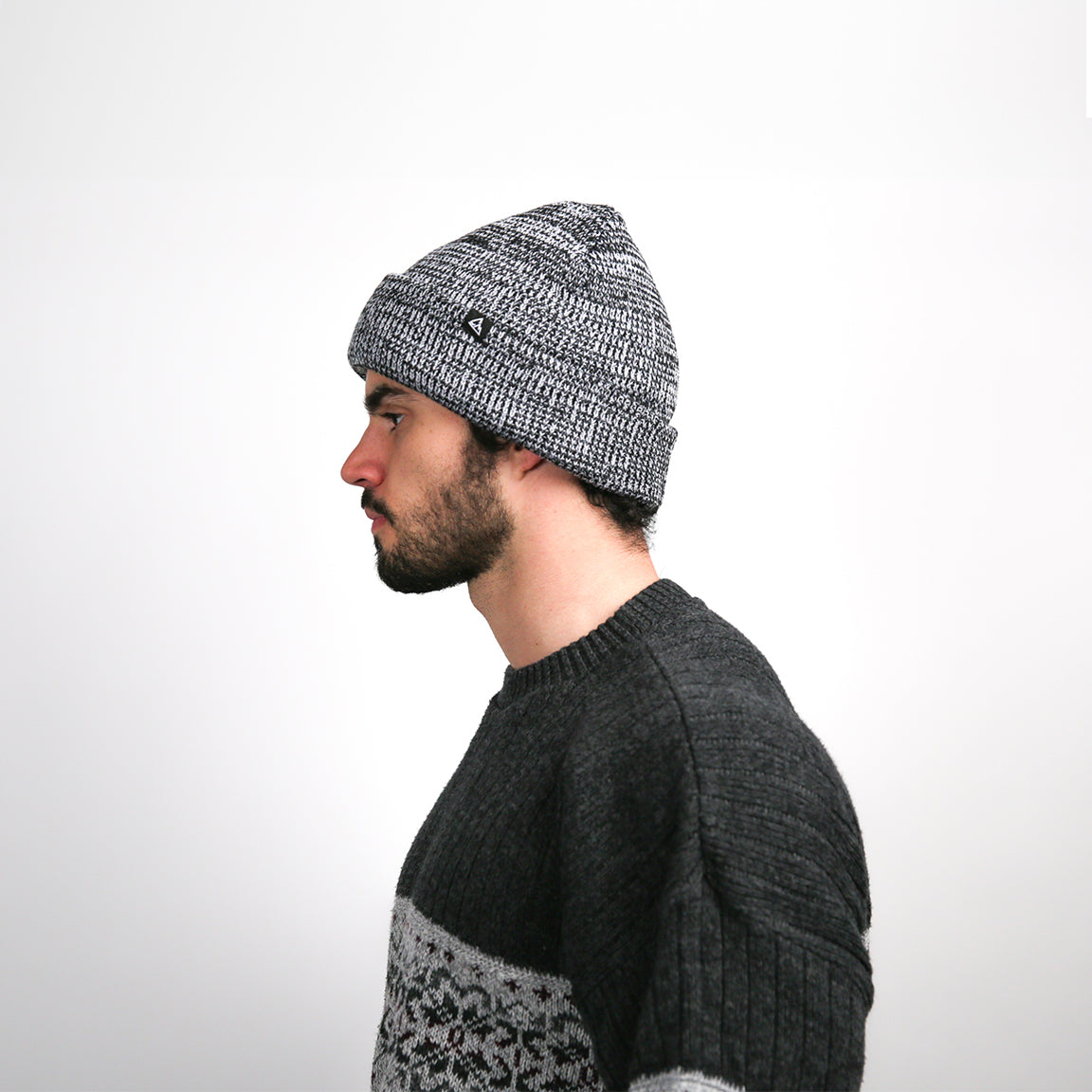The two-tone grey knit beanie is viewed from the side, highlighting the intricate texture and the beanie's easygoing drape, complemented by a small black logo patch on the brim.
