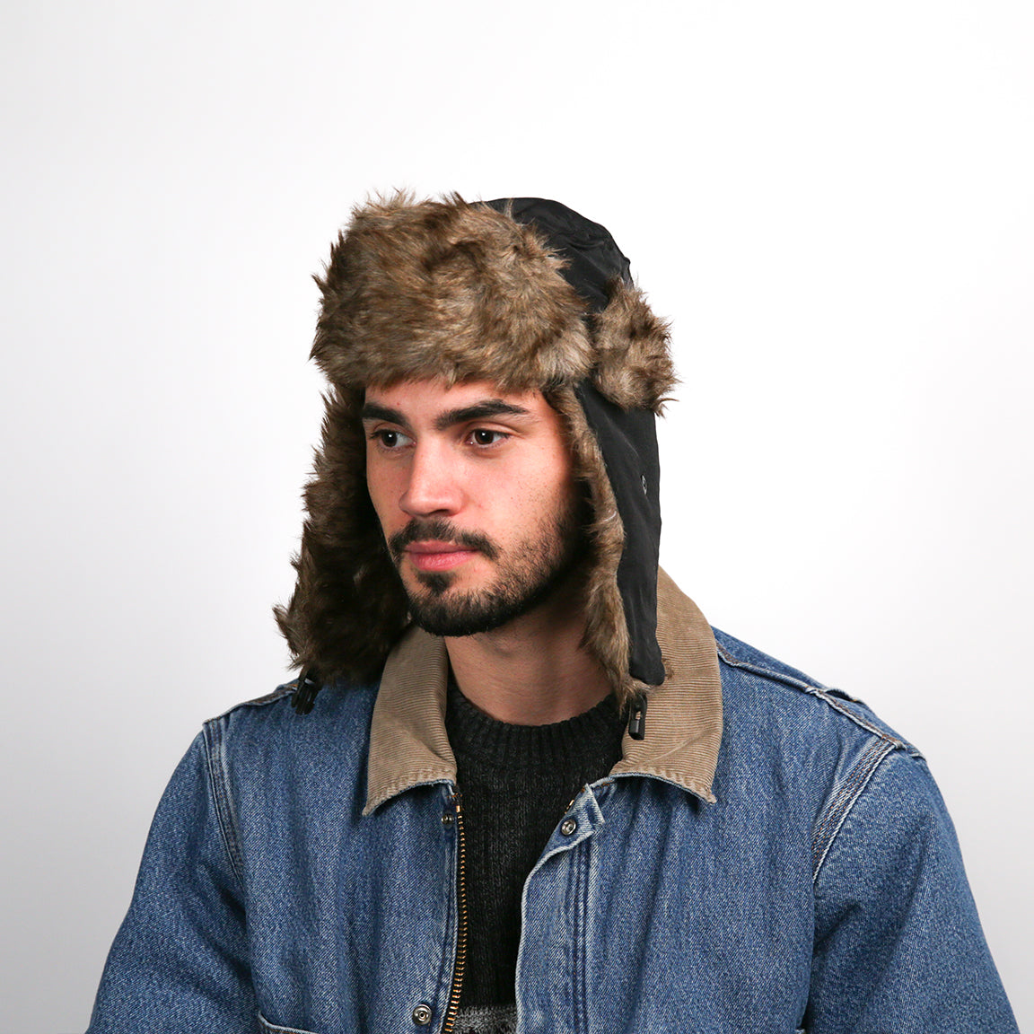 A person is shown wearing an aviator hat with a brown faux fur lining and black outer shell. The hat features ear flaps that can be secured up or down and a buttoned chin strap.