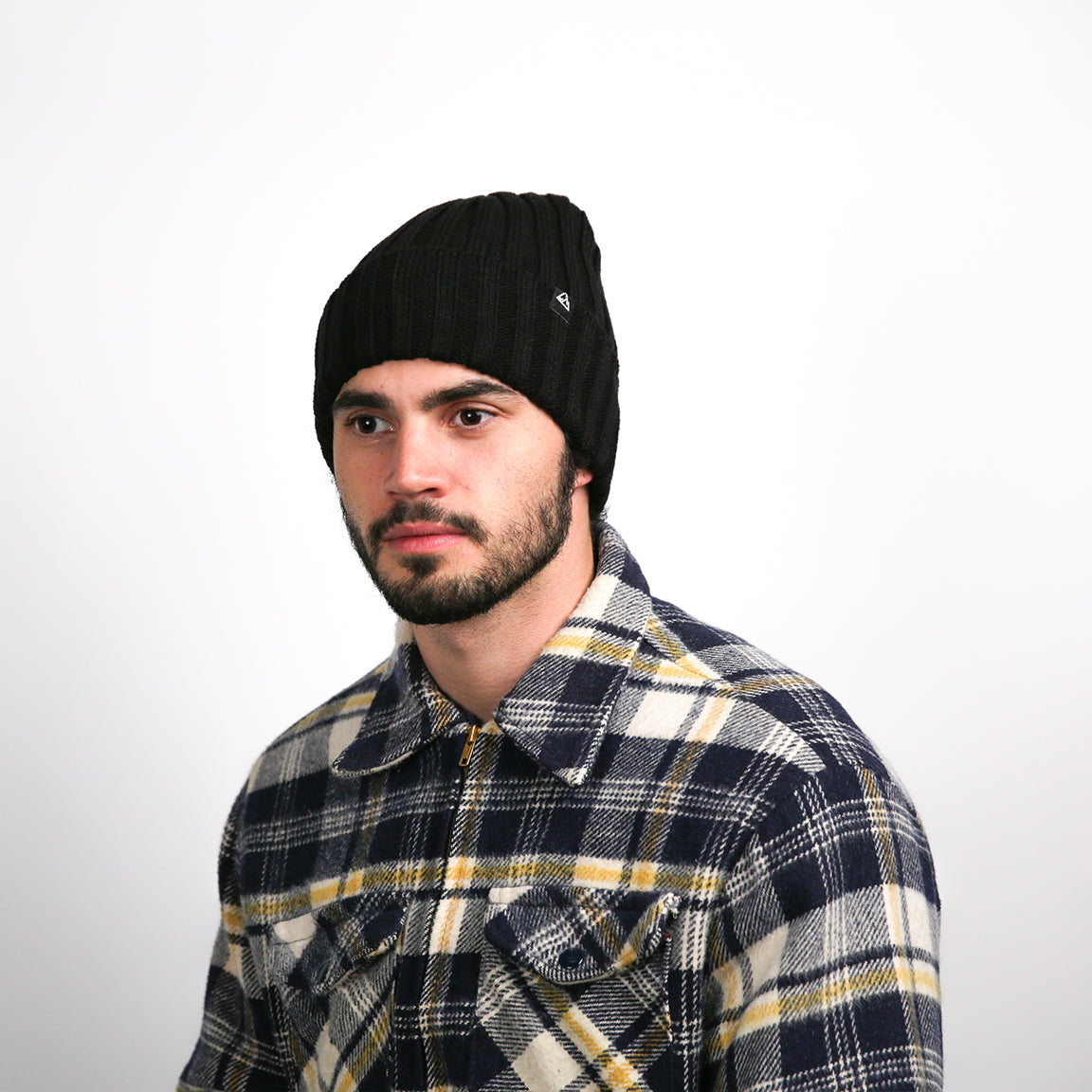 A person is shown wearing a ribbed, black beanie that fits snugly around the head. The beanie has a textured pattern, with a small, black triangular logo on the fold near the forehead. The person is also wearing a plaid shirt with navy blue, white, and yellow checks