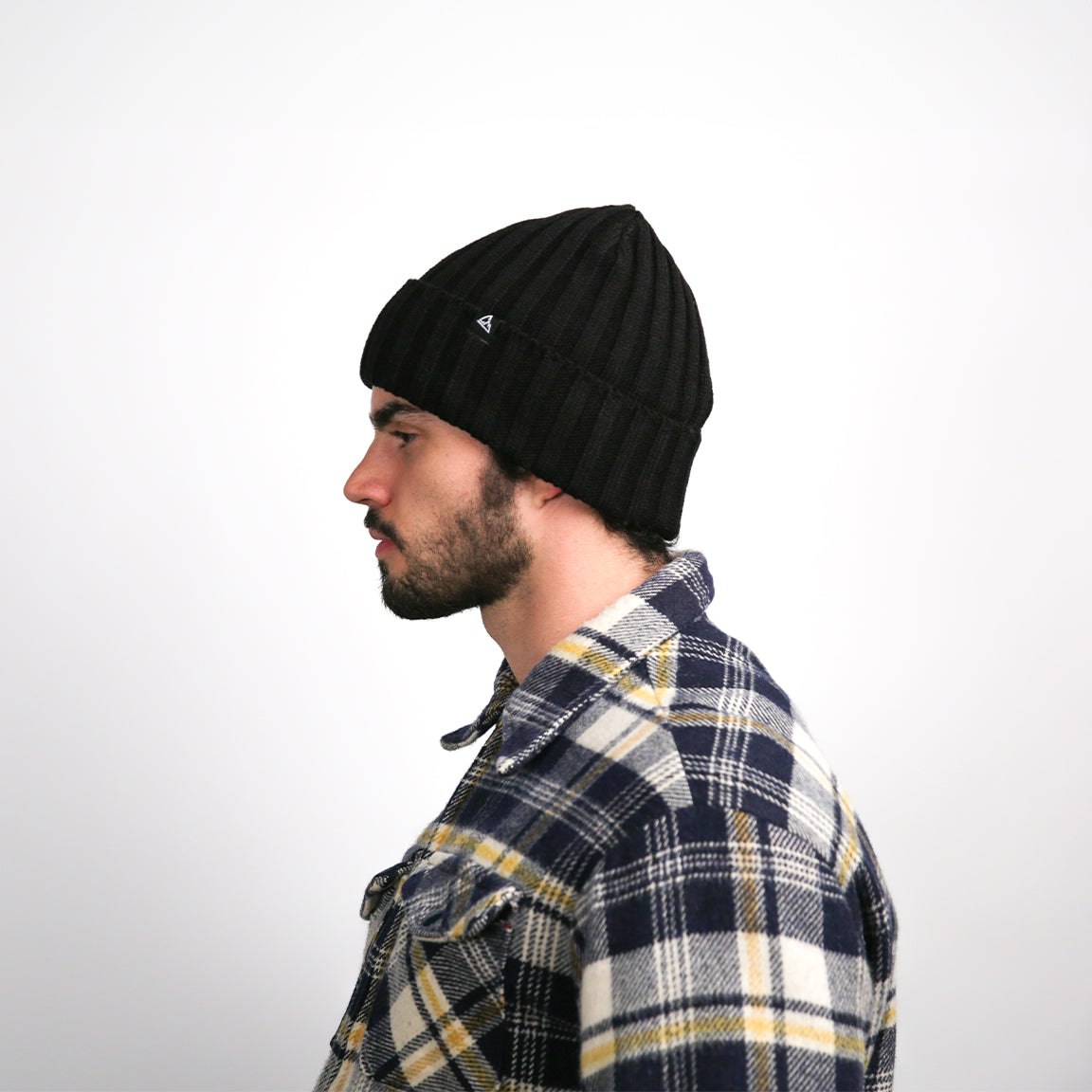 A side view of a person shows the profile of the black beanie, where the ribbed texture and the logo are visible. The beanie covers the ears and shapes to the head, ending with a slight rounded excess at the top, creating a comfortable fit. The plaid shirt's pattern is also visible from this angle.
