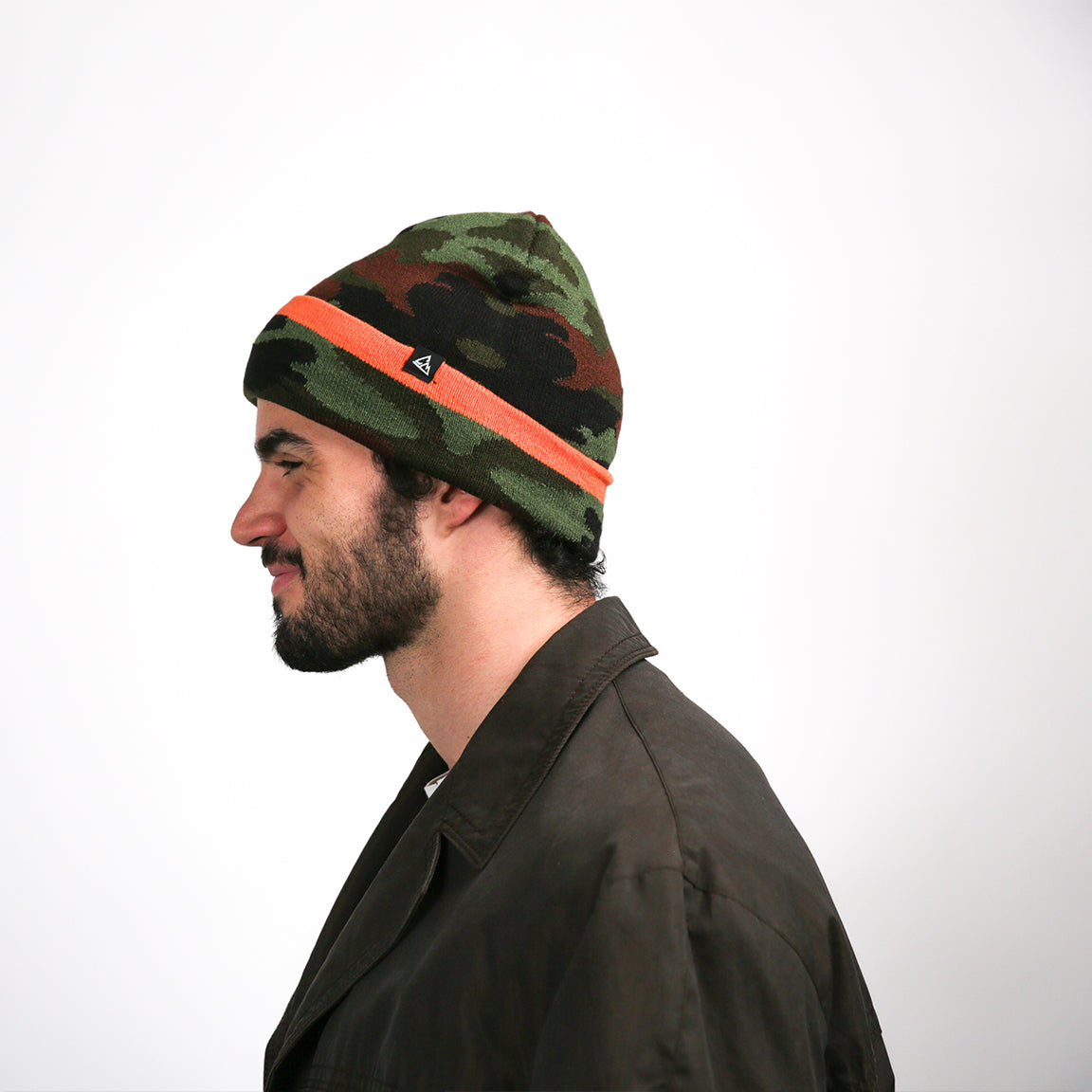 A side profile view of the same camouflage-patterned beanie. The orange stripe and black logo are visible on the side, contrasting with the camo design. The beanie maintains its slouchy shape towards the back.