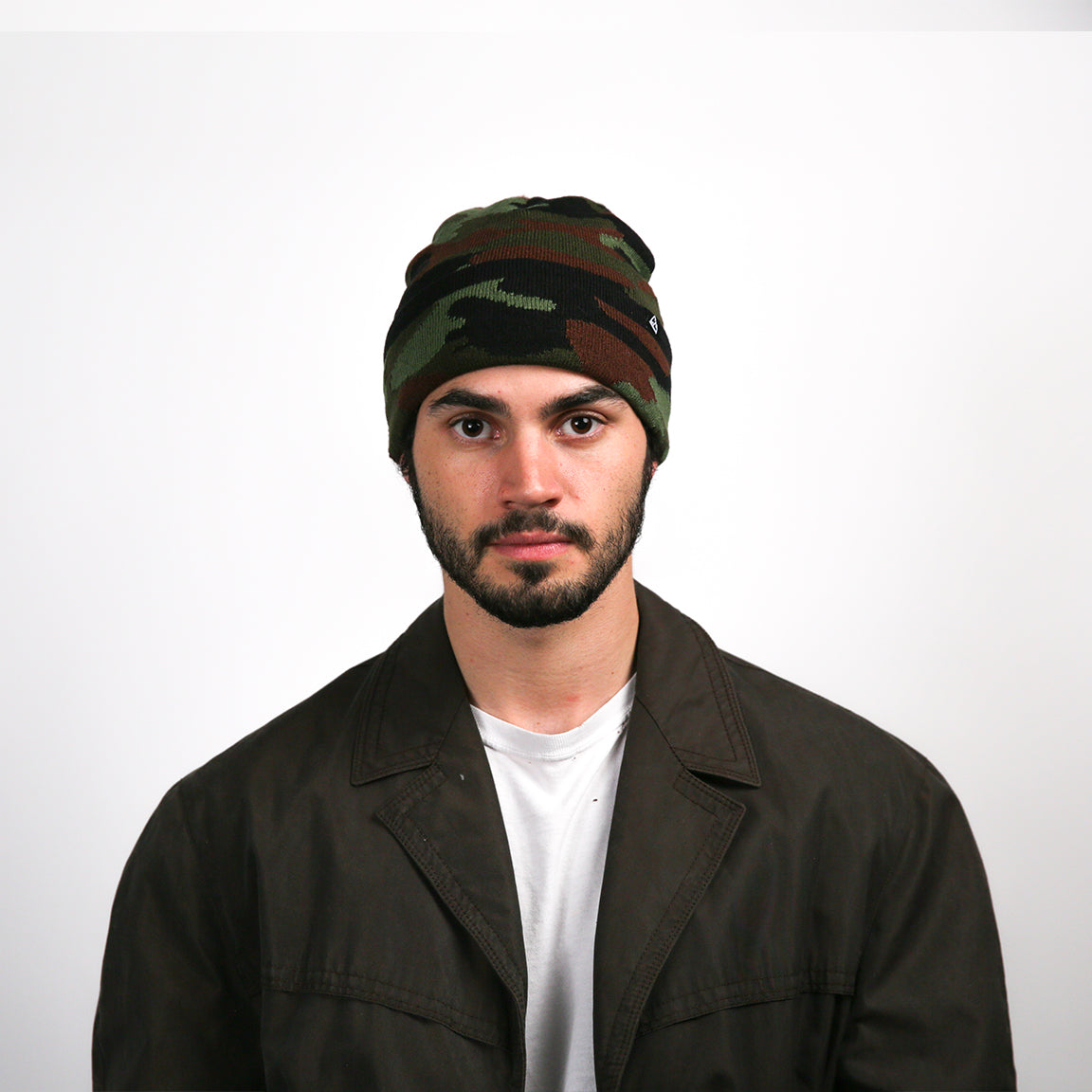 The beanie presented is knitted with a camouflage pattern that blends dark green, brown, and black tones. It is accented by a black stripe near the bottom edge, where a small black logo is also visible. The beanie is designed to fit closely to the head with a slight slouch at the crown.