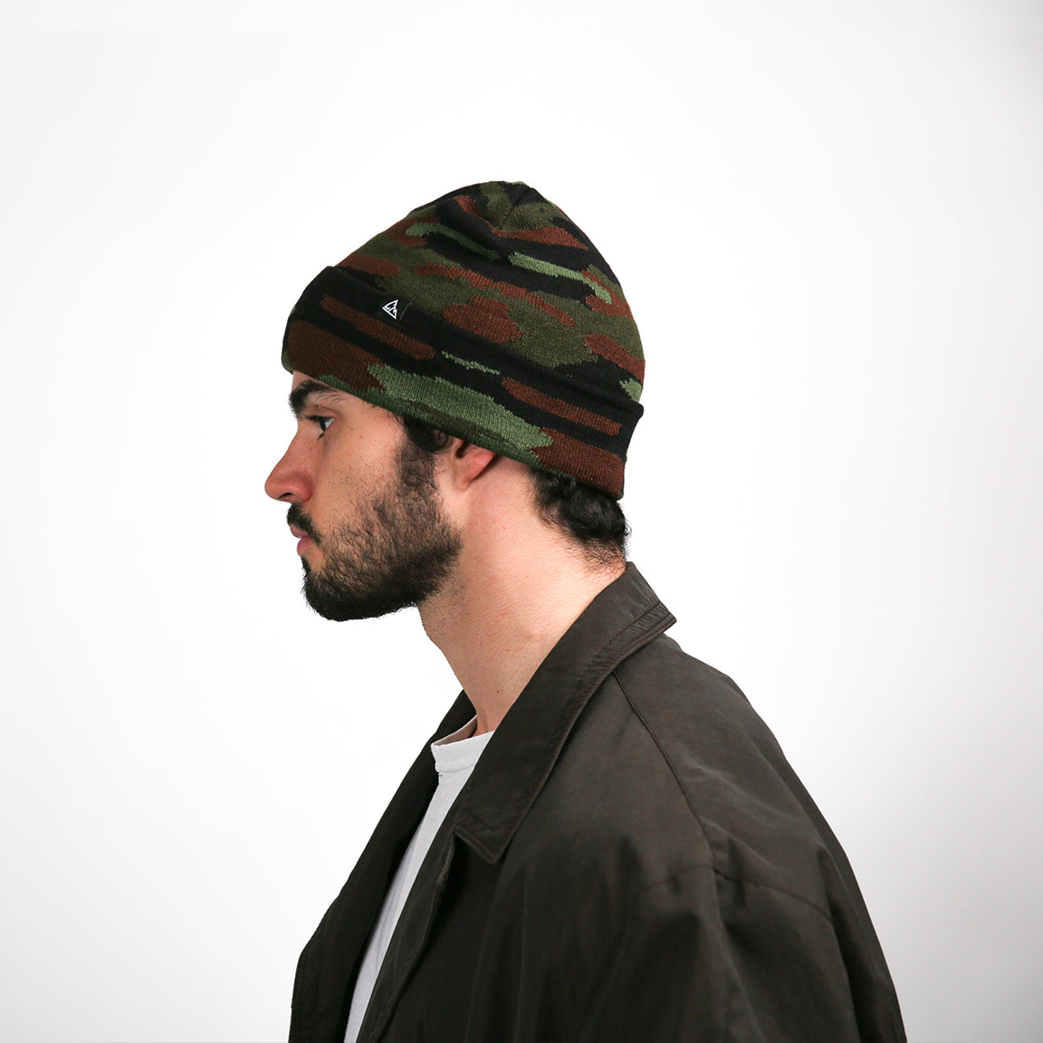 This side view shows the same beanie, where the black stripe encircles the hat, contrasting with the camouflage pattern. The black logo is positioned just above the stripe. The beanie conforms to the head's shape, with a gentle slouch evident at the back.