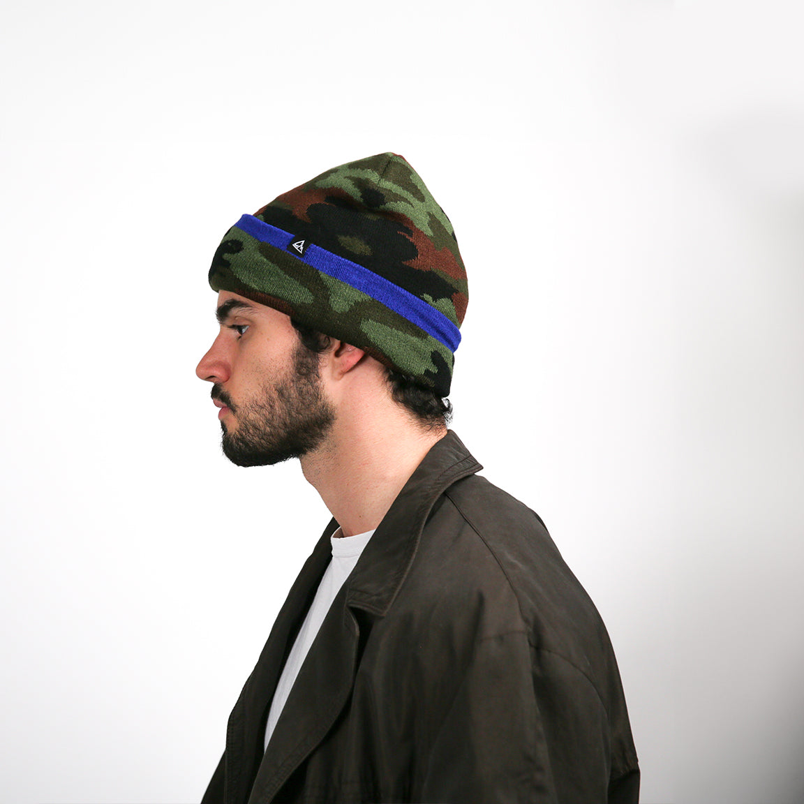A beanie is viewed from the side. The bright blue stripe runs horizontally around the head, standing out against the dark camouflage pattern. The logo is also visible on the side. The beanie's fabric drapes softly to one side, maintaining its relaxed, slouchy look.