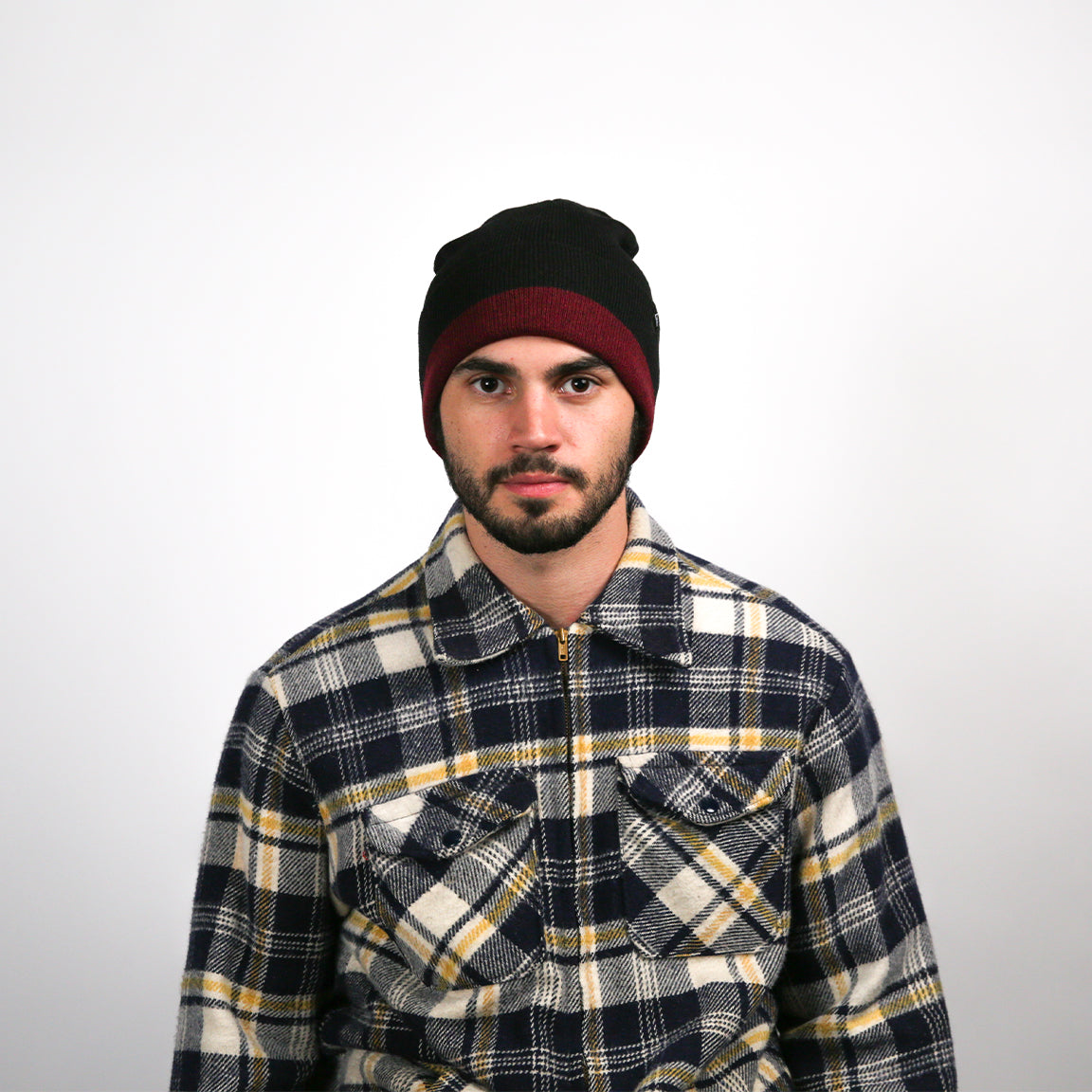 A man with a medium complexion stands directly facing the camera. He has dark eyes, a neatly trimmed beard, and short hair, which is mostly covered by a snug-fitting beanie that is black with a red rim. He's wearing a cozy flannel shirt with a plaid pattern combining navy blue, white, and mustard yellow, featuring buttoned chest pockets.