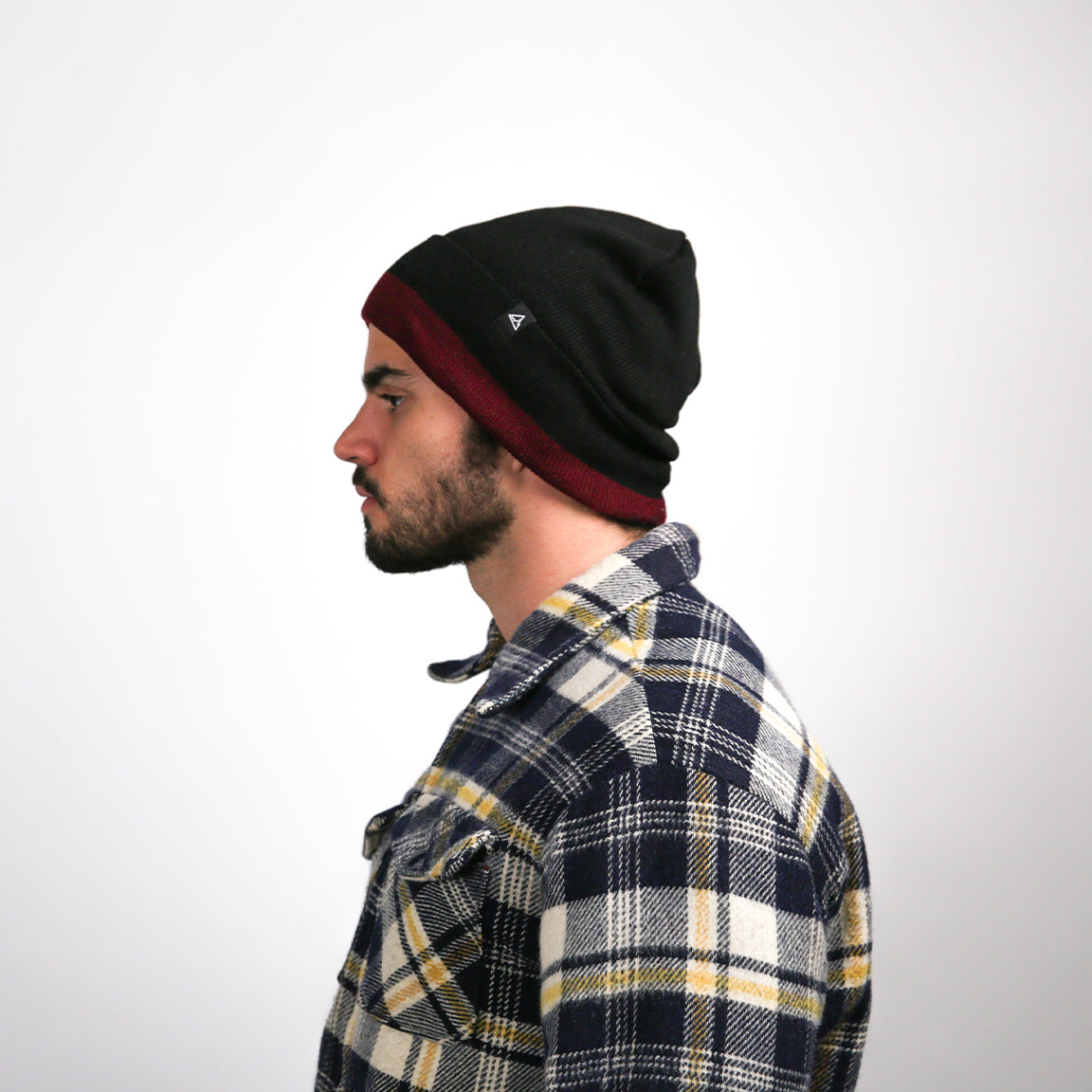 A side profile shows the man turned to his right, presenting the shape of his beanie, which has a small triangular logo on the fold. His facial features are sharp, and his expression is neutral. The plaid shirt's collar is neatly turned down, and the fabric appears soft and thick, suitable for cool weather.