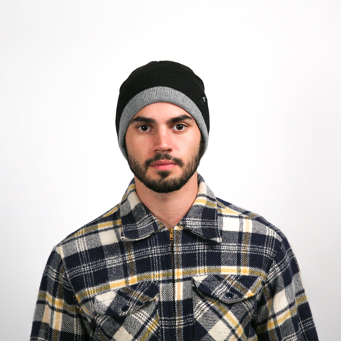A man with a medium complexion and a focused gaze is facing the camera. The beanie he wears is black with a wide, heather grey band at the bottom. His flannel shirt is a combination of navy blue, white, and a bright mustard yellow in a plaid pattern. The shirt's material appears thick and warm.