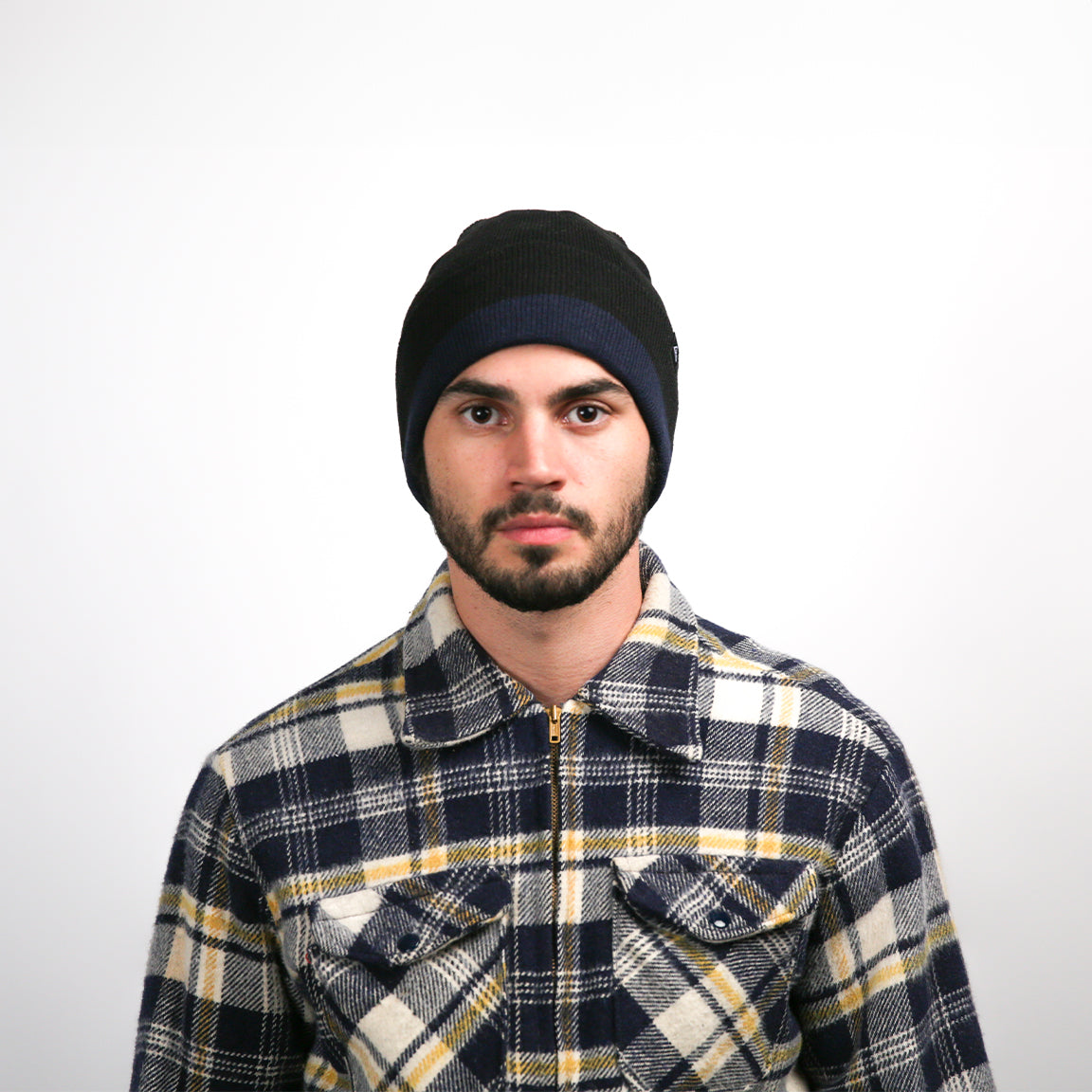 The man is directly facing the viewer with a neutral expression. His beanie is black with a navy blue band around the edge. The soft, thick texture of his plaid flannel shirt, featuring navy blue, white, and mustard yellow squares, is visible. The fabric has a slight sheen, indicating its thickness and warmth.