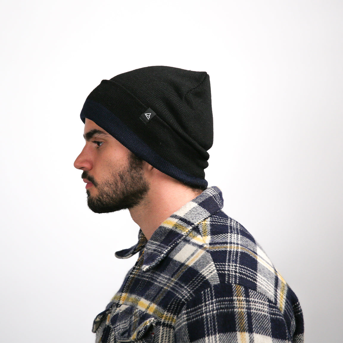 Seen from the side, the man’s beanie is bunched at the back, suggesting a relaxed fit. The shirt's plaid pattern, with large squares, is noticeable, and the shirt's thick fabric folds naturally at the elbow. His profile is calm, with a straight nose and a slightly open mouth.