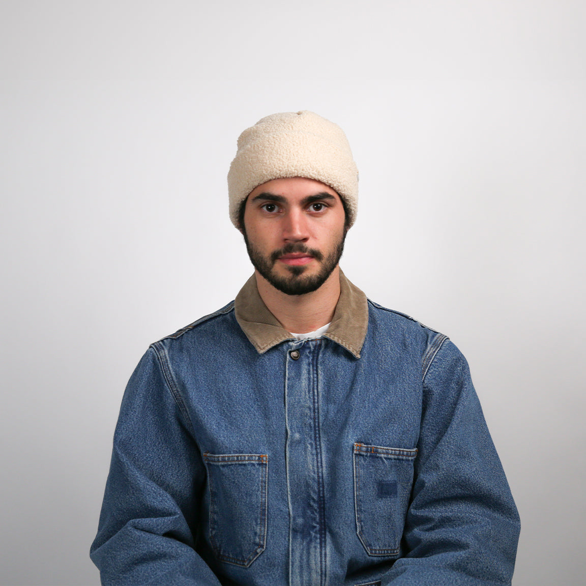 A person facing forward, wearing a cream-colored fleece beanie. The beanie is textured, soft to the touch, and has a rolled-up brim, which adds to the thickness around the head. The fit is relaxed, with a bit of room at the top, which gives it a slightly slouchy look. The person is dressed in a blue denim jacket with a contrasting tan collar.