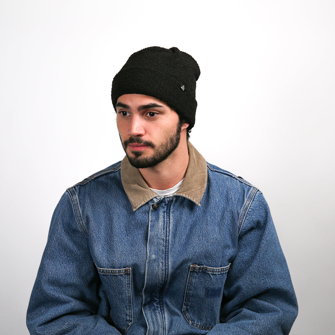 A person is wearing a black fleece beanie with a similar texture and rolled-up brim as the first. A small black triangular logo is attached near the edge. The beanie fits snugly around the head with a gentle slouch at the back. The denim jacket with the tan collar is again part of the attire.