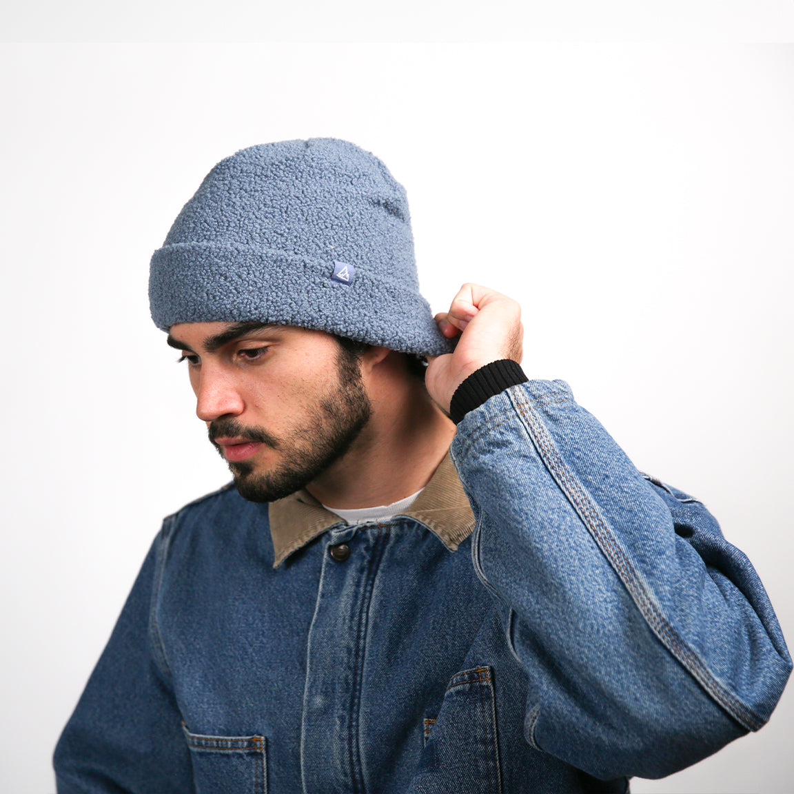 A person wearing a beanie that is a soft blue fleece with a rolled-up brim and a small blue triangular logo. The person is adjusting the beanie at the back, indicating a flexible and comfortable fit. The slouchy top suggests a relaxed style, complementing the casual denim jacket with its tan collar.