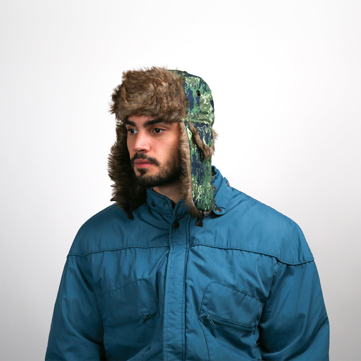 Pictured is a person wearing an aviator hat with a green camo pattern and brown faux fur lining. The design includes functional ear flaps and a buttoned chin strap, suited for outdoor activities.