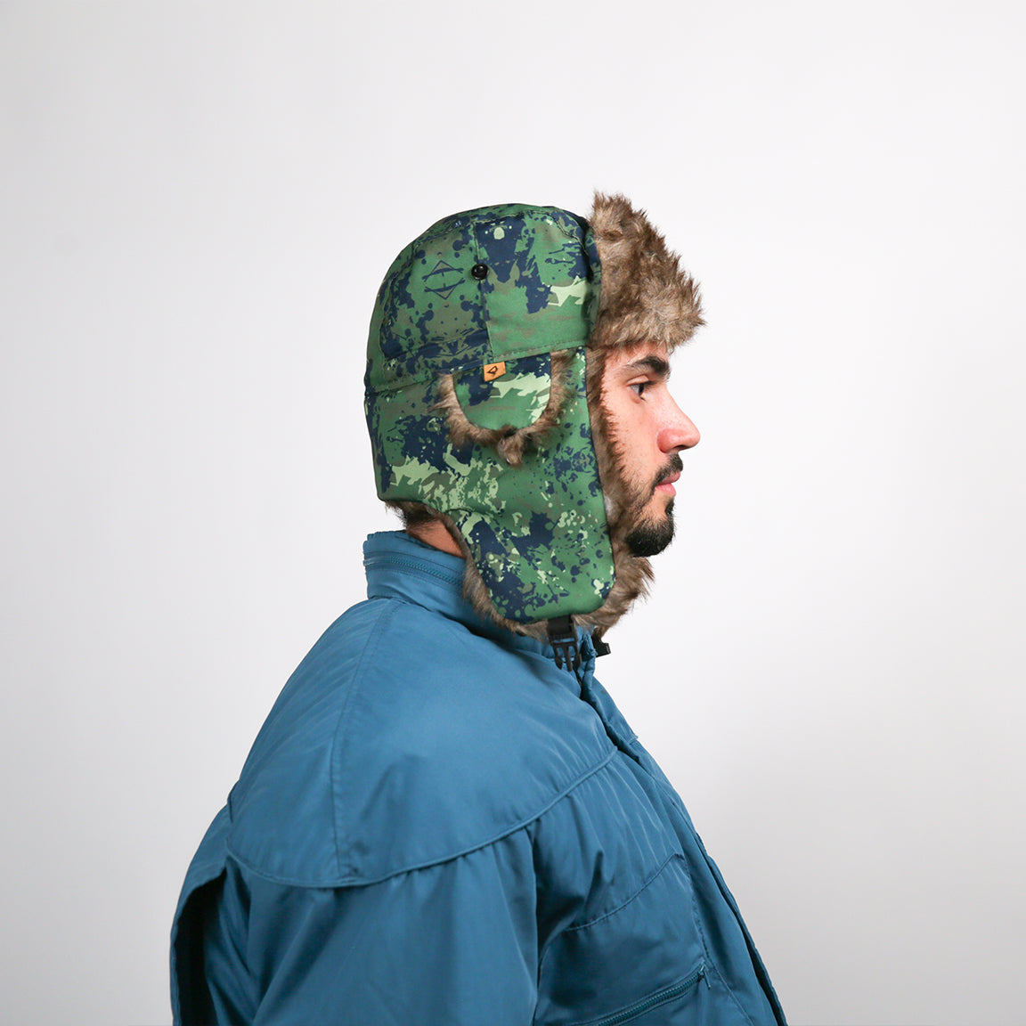 Pictured is a person wearing an aviator hat with a green camo pattern and brown faux fur lining. The design includes functional ear flaps and a buttoned chin strap, suited for outdoor activities.
