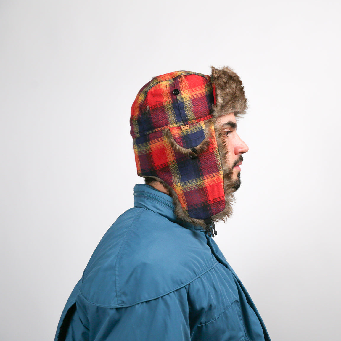 The person models an aviator hat featuring a vibrant red plaid pattern with a brown faux fur lining. The hat's design includes ear flaps and a chin strap for secure and adjustable fitting.