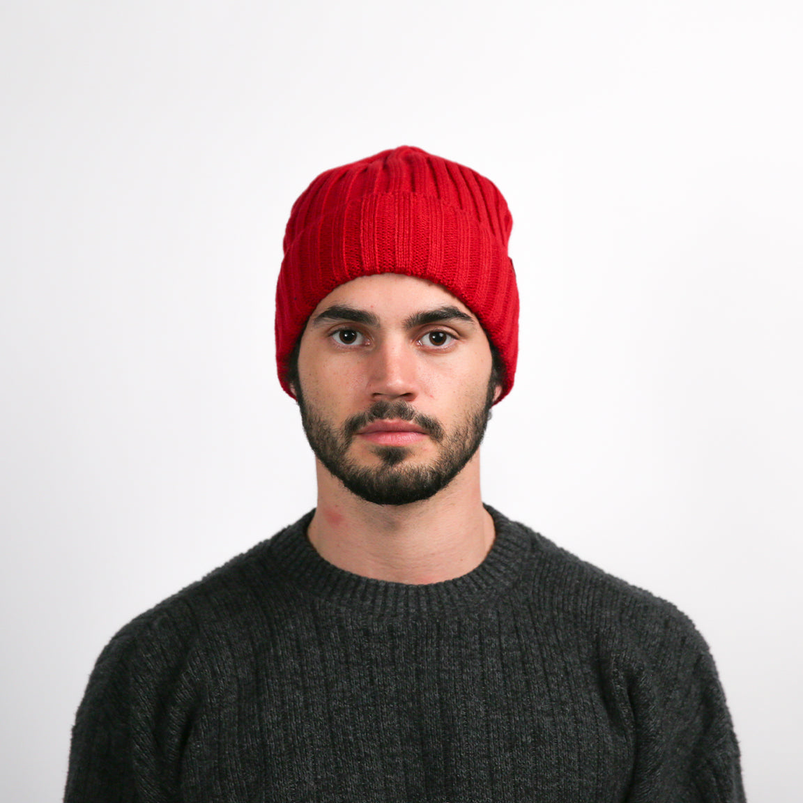 A person facing forward, wearing a vibrant red, ribbed beanie that sits snugly on the head. The beanie is folded at the brim where a small, white triangular logo is visible. The beanie has a relaxed fit at the top, suggesting a slight slouch. The person is also wearing a charcoal grey knit sweater with a prominent weave pattern.
