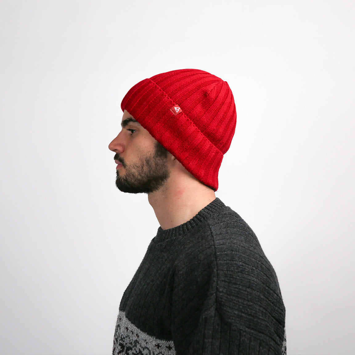 Displayed in a side profile, the same red beanie shows the ribbed texture and the fold at the brim with the white logo. It covers the ears and conforms to the head's shape, with the material gathering at the top for a casual, slouchy appearance. The texture of the charcoal grey sweater is also visible from this angle.