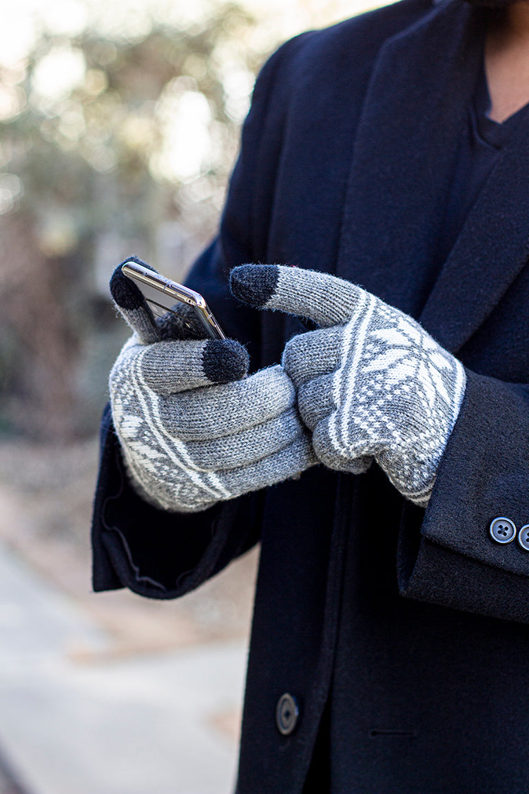A person in a navy coat wears gray touchscreen gloves with white patterns, using a smartphone. The gloves' fingertips are smartphone-friendly.